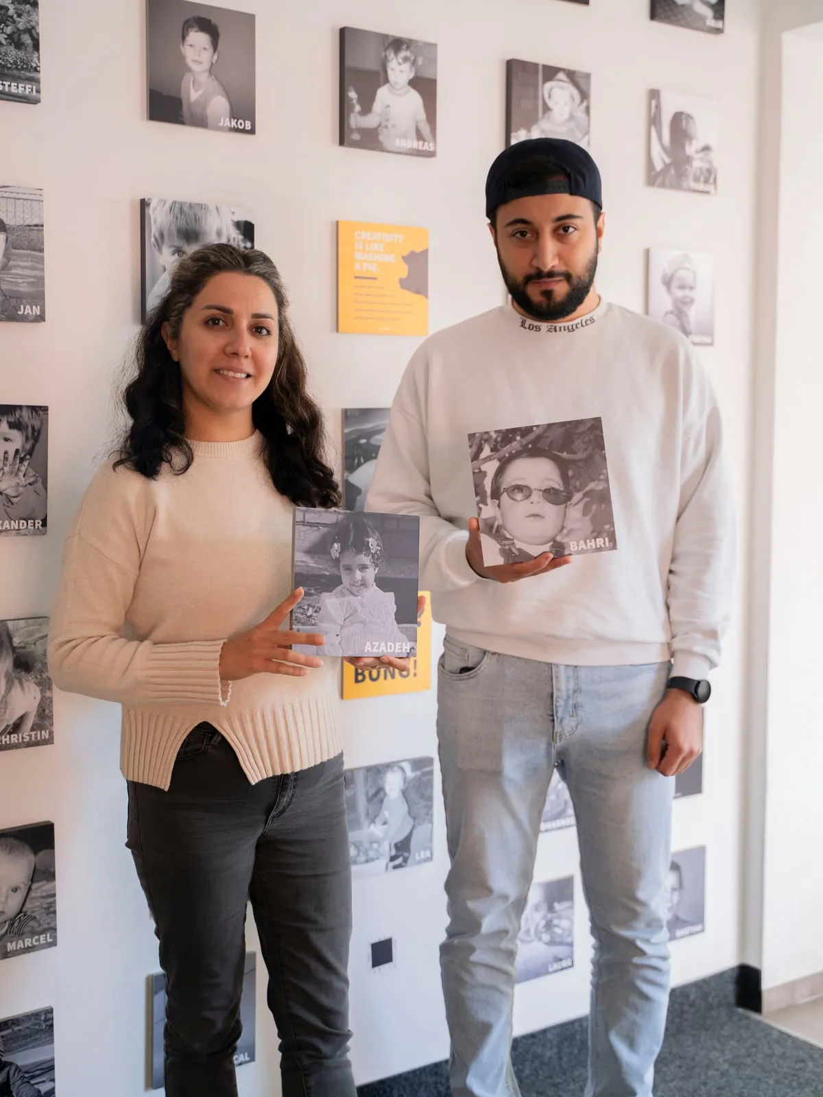 Azadeh und Bahri stehen vor der Wand mit den Kinderfotos des C3-Teams und halten ihr eigenes Kinderfoto in den Händen