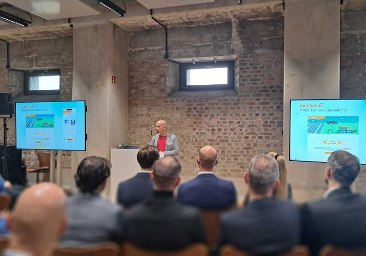 Christian Reisnecker präsentiert VEDA-Markenstrategie bei der Mitgliederversammlung in München Christian Reisnecker steht an einem Rednerpult im Bergson Kunstkraftwerk München und präsentiert das neue Web- und Markenprojekt für die Vereinigung der deutschen Autohöfe (VEDA). Zwei große Bildschirme flankieren die Bühne, das Publikum verfolgt aufmerksam die Präsentation.