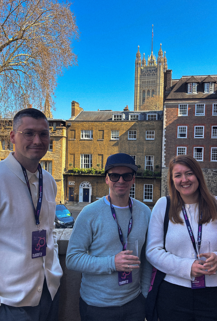 Alexander Schirmer, Christian Reisnecker und Anja Meixner lächeln für ein Porträtfoto auf einem Balkon. Alle drei tragen lilafarbene Lanyards der „Marketing Meetup“-Konferenz. Im Hintergrund erstreckt sich die Londoner Architektur mit historischen Backsteingebäuden und der markanten Spitze des Victoria Tower vom Palace of Westminster unter einem klaren blauen Himmel.
